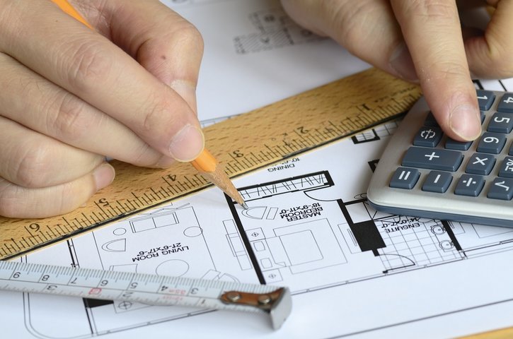 What Is A Pre Demolition Audit? This image shows building plans, a calculator and ruler with a hand marking the pln in pencil