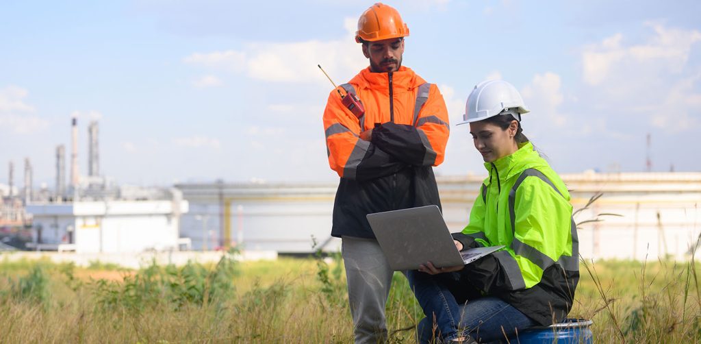 The Role of Environmental Consultancy in Modern Construction this image shows environmental engineers on a lap top in a field with an oil refinery in the background
