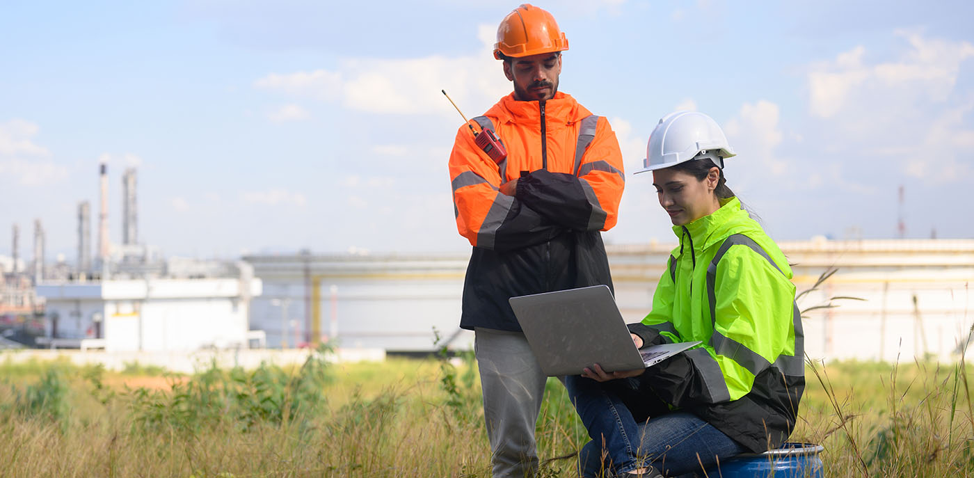 The Role of Environmental Consultancy in Modern Construction this image shows environmental engineers on a lap top in a field with an oil refinery in the background